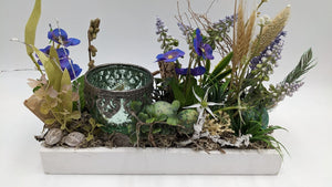 Ostergesteck in weißer Holzschale mit grünem Teelichtglas, lila Blüten, Hyazinthen, Ähren und gesprenkelten Eiern