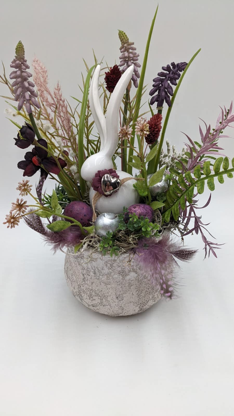 Frühlingsgesteck von vorn: Hase mit Silberherz, Farn, Gräser, Beeren-Eier und Federn in Lila und Weiß