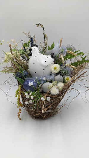 Ostergesteck im Reisignest mit weißem Keramikhuhn, blauen und graugrünen Eiern, Bellis und Gräsern