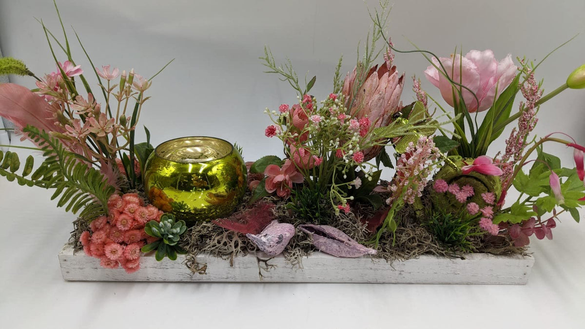 Längliches Frühlingsgesteck in Rosa und Grün auf weißem Holztablett mit grünem Teelichtglas, Tulpe und Federn