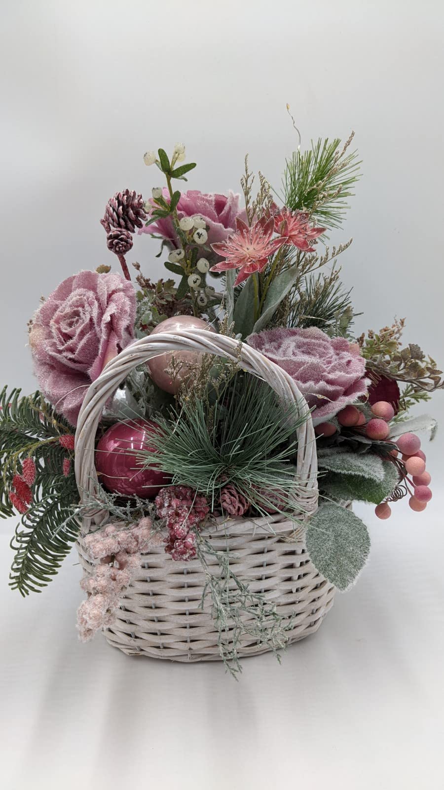 Wintergesteck im weißen Korb mit altrosa Rosen, Kugeln, Tanne und Zapfen, dazwischen feine LED-Lichterkette