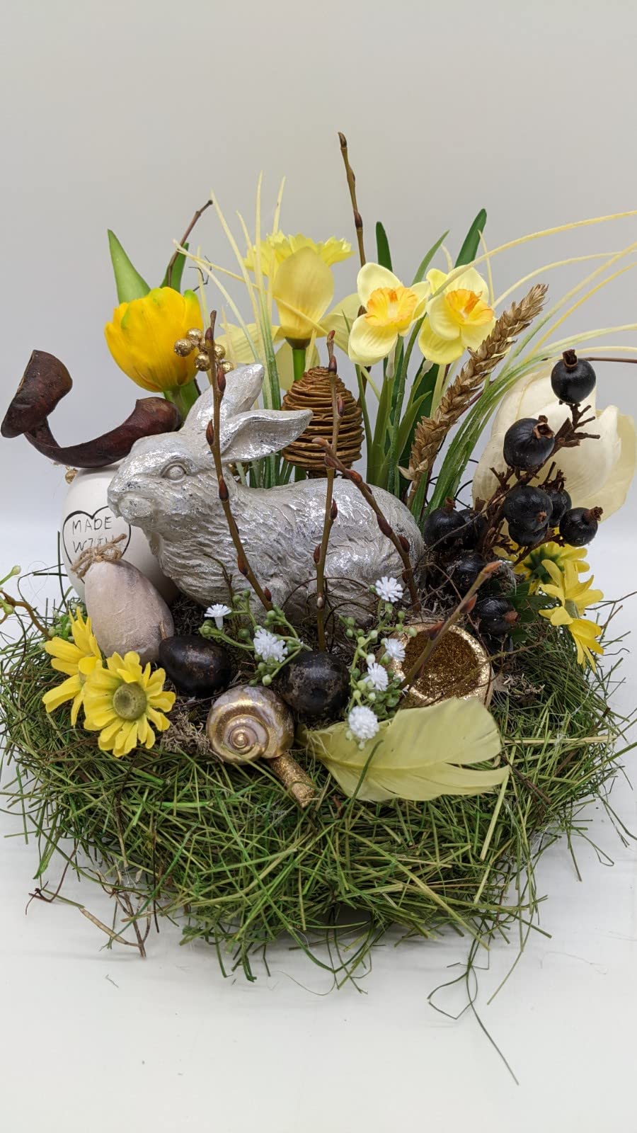 Ostergesteck von vorn: silberner Hase, gelbe Tulpe, Narzissen, dunkle Beeren und Eier im grünen Nest