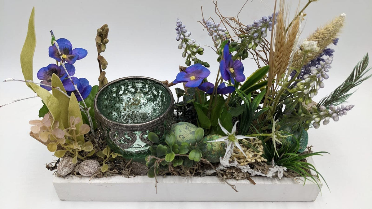 Frühlingsgesteck von vorn: Teelichtglas, violette Blüten, hellblaue Hyazinthen, Gräser, Moos und grüne Ostereier