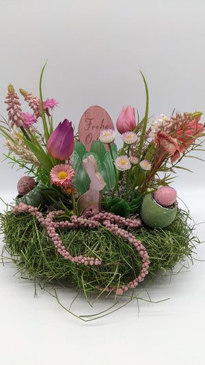 Ostergesteck auf Grasnest mit rosa Tulpen, Bellis, Holzhase und gesprenkelten Eiern in grünen Eierschalen