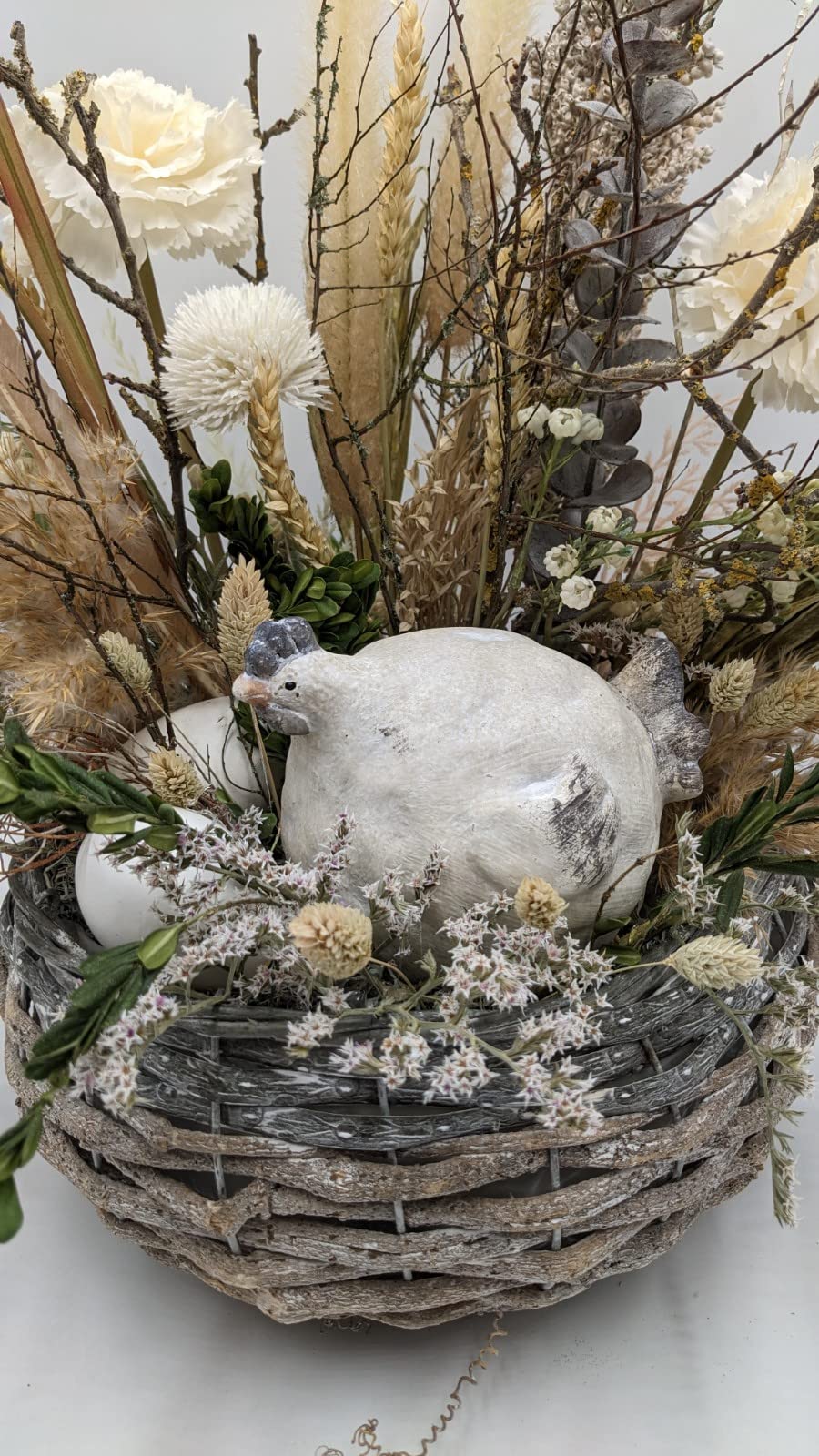 Nahaufnahme des weißen Huhns im Nest zwischen weißen Eiern, Ähren, Buchsblättern und zartrosa Trockenblüten