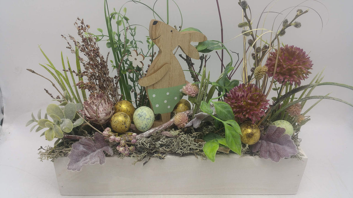 Ostergesteck im weißen Holzkasten mit Holzhäsin, goldenen Eiern, lila Allium und Gräsern