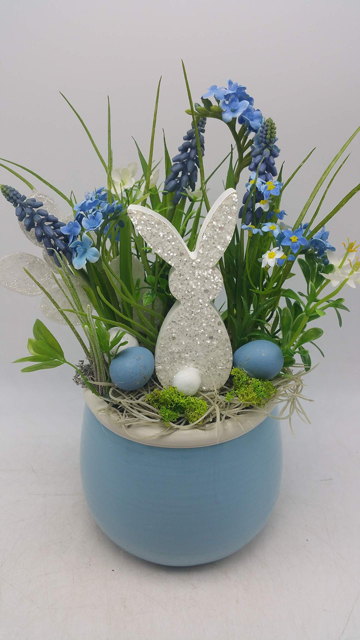 Ostergesteck mit weißem Glitzerhasen, blauen Eiern, Traubenhyazinthen und Gräsern in hellblauem Keramiktopf