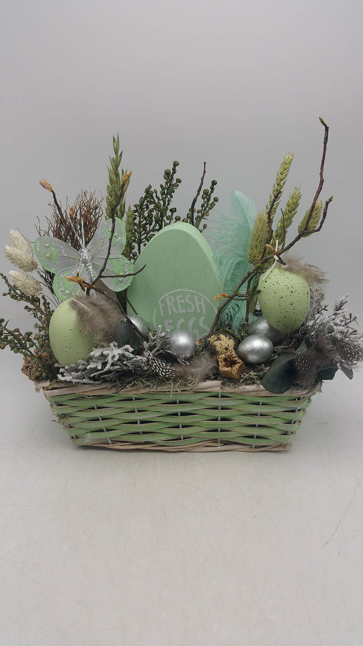 Ostergesteck von vorne: flacher Weidenkorb in Mint mit grünen und silbernen Eiern, Ähren und Zweigen