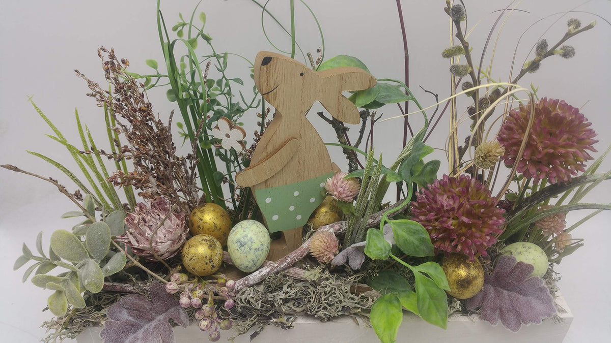 Nahaufnahme des Ostergestecks: Häsin mit grüner Hose, gesprenkelte Eier, Moos und rosa Blüten