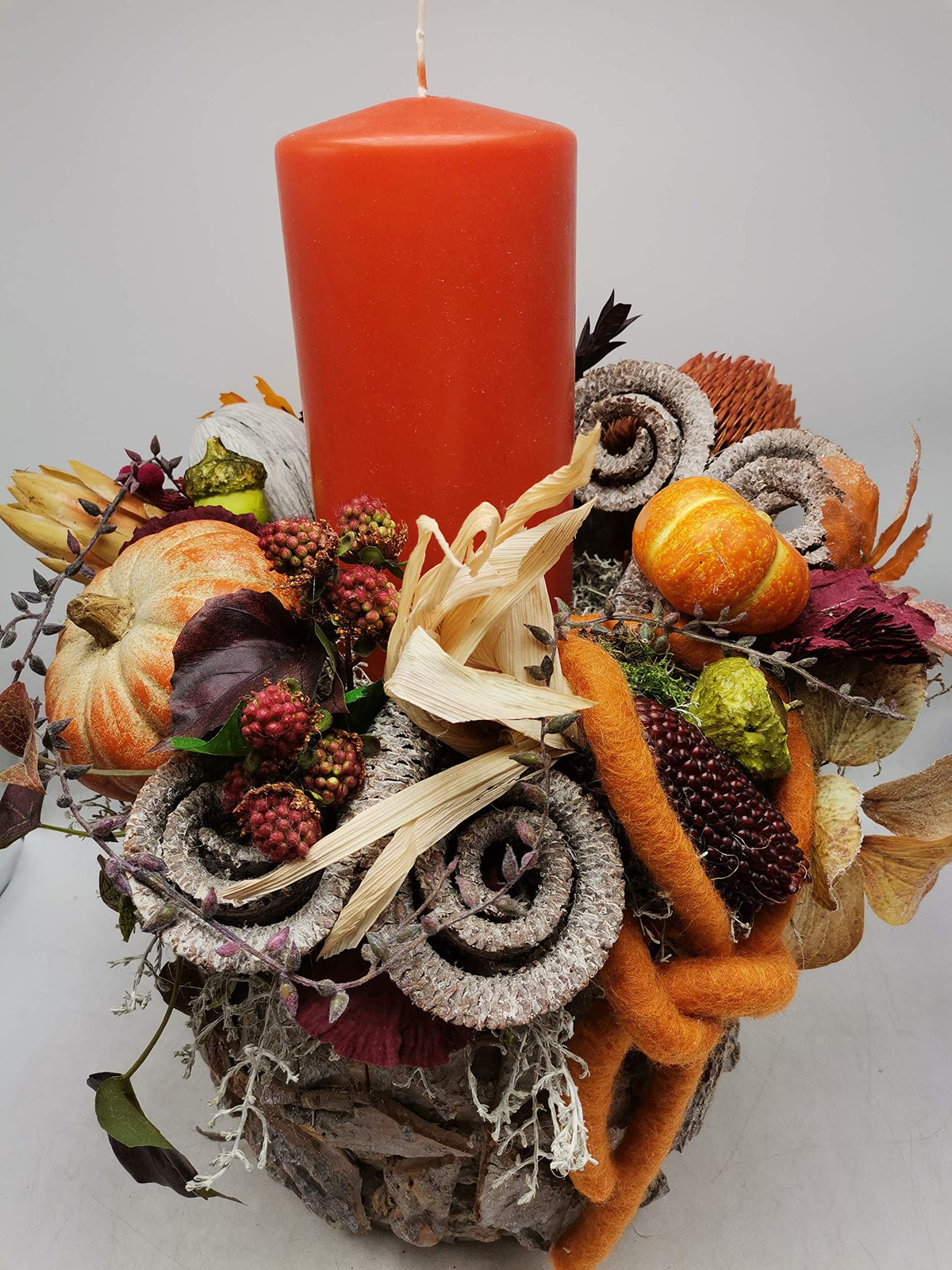 Herbstgesteck von oben: orange Kerze, Kürbis in Orange-Creme, roter Ziermais, Beeren und gerollte Rinde