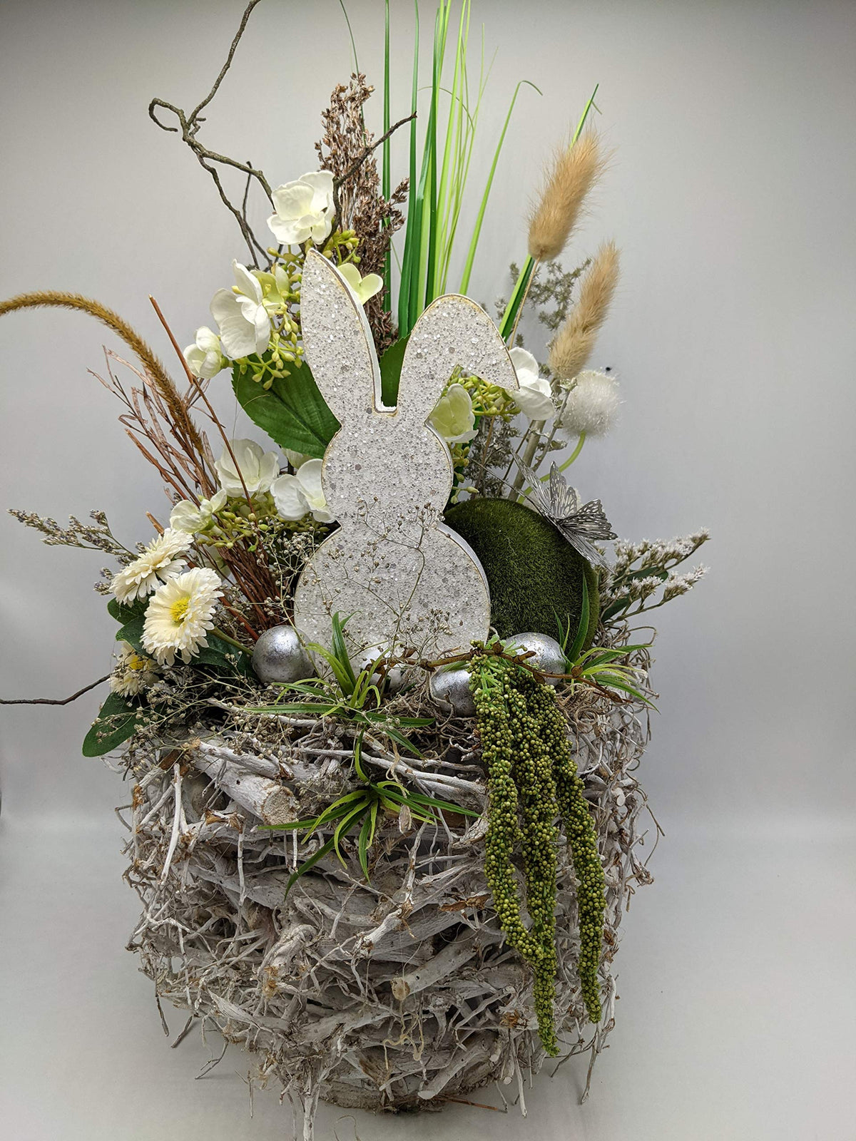 Ostergesteck auf weißem Zweiggeflecht mit Holzhase, Bellis, Hortensien, Gräsern und silbernen Eiern