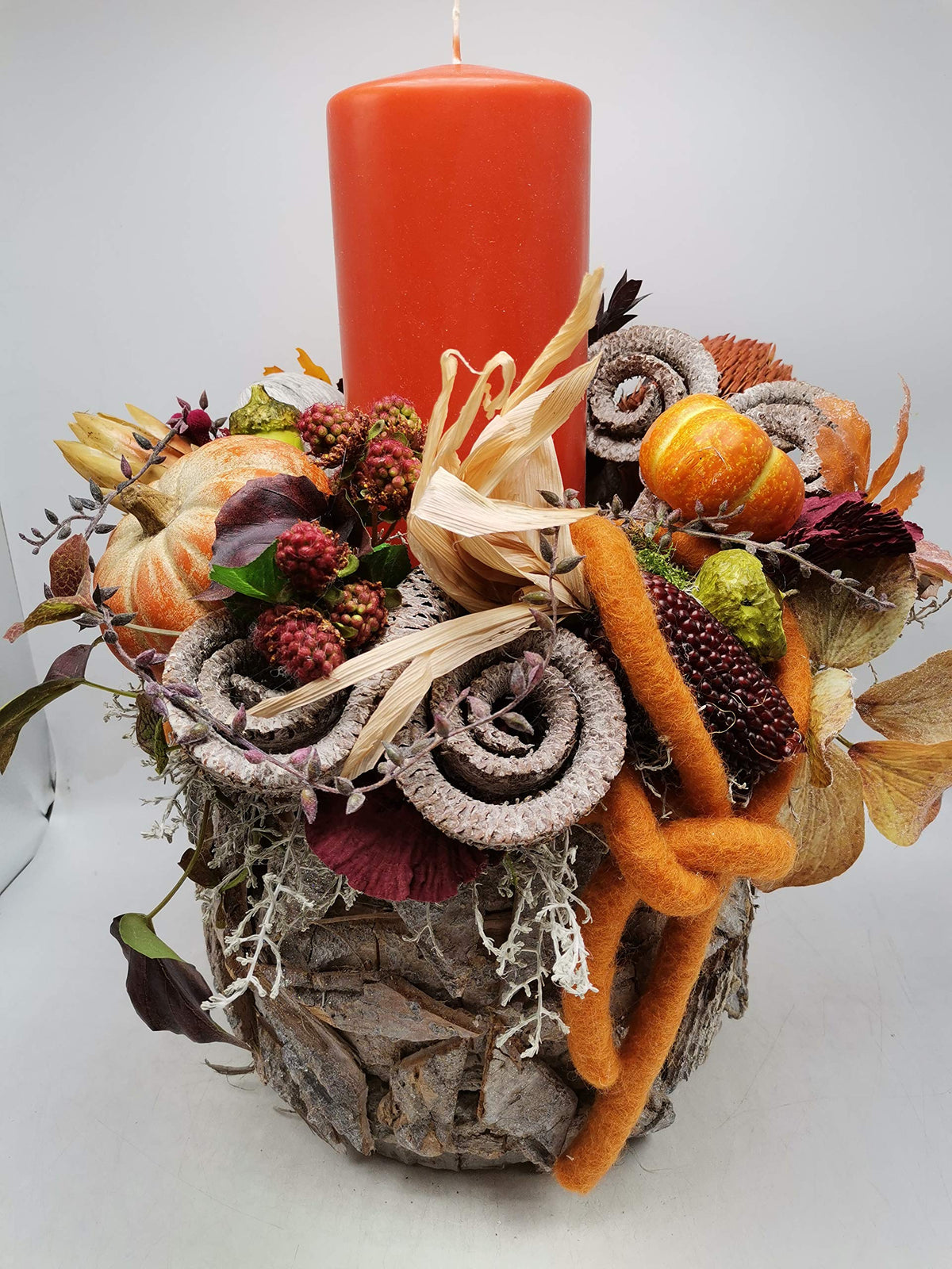 Herbstgesteck mit orangefarbener Stumpenkerze, Kürbissen, Ziermais und Beeren in Rindentopf, Ansicht von vorn