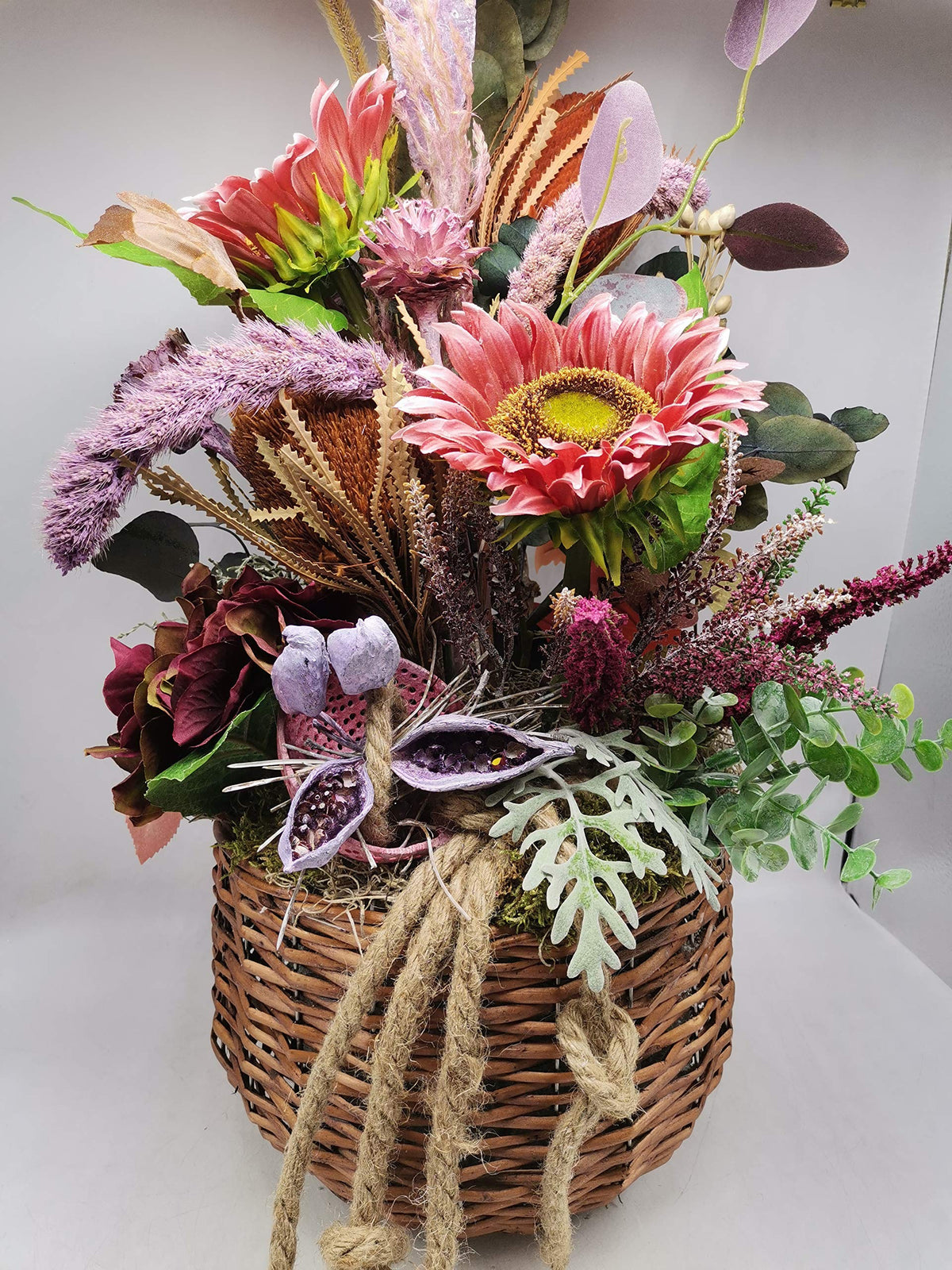 Herbstgesteck im Weidenkorb mit rosa Sonnenblumen, Banksia, Heide, lila Schoten und Eukalyptus