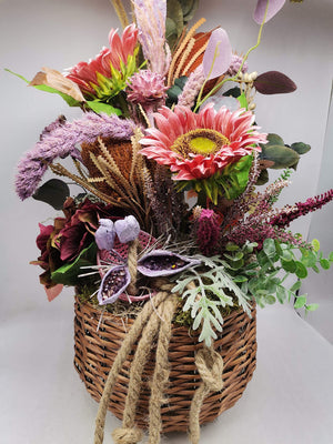 Herbstgesteck im Weidenkorb mit rosa Sonnenblumen, Banksia, Heide, lila Schoten und Eukalyptus