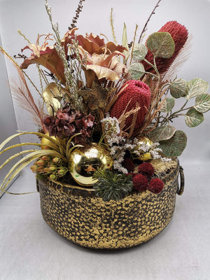 Herbstgesteck mit roten Banksia, kupferfarbenen Trompetenblumen und goldenen Kugeln in gold-schwarzer Metallschale